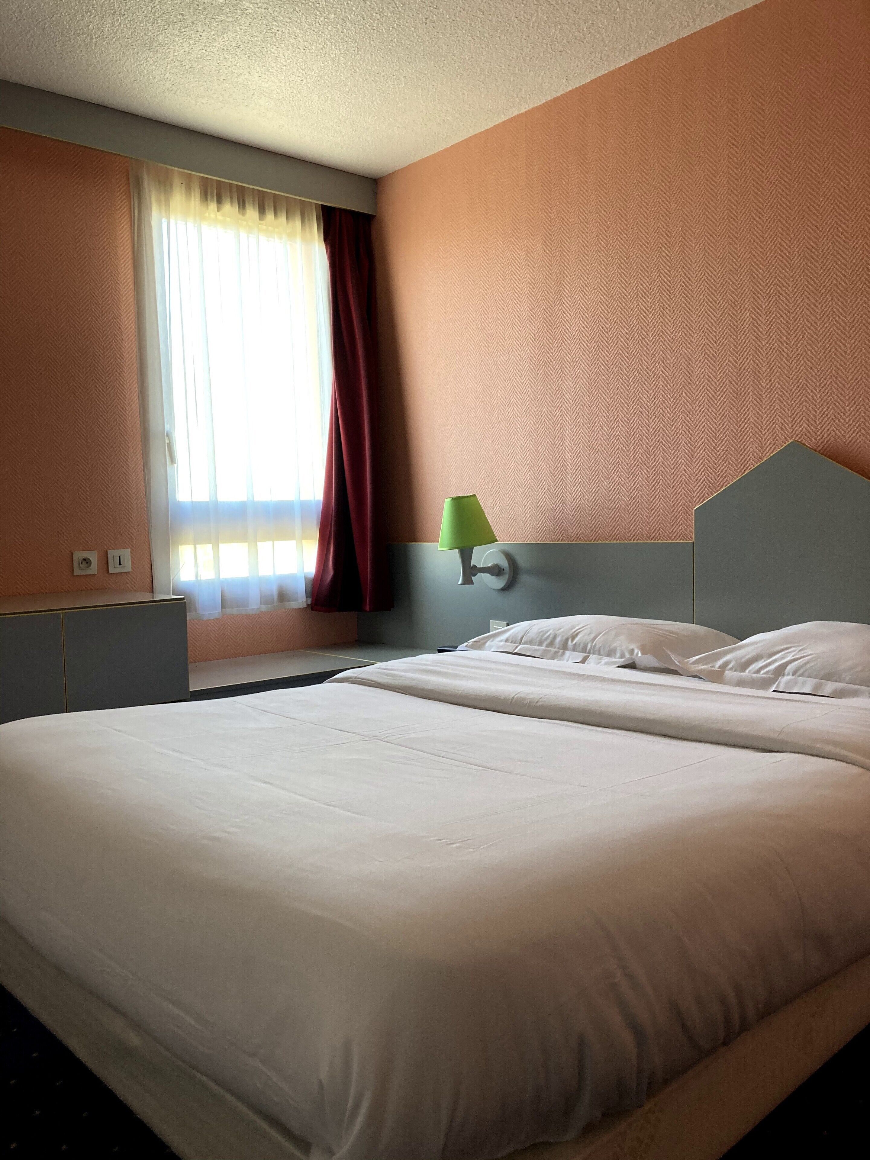 A hotel room with a large bed, a nightstand, a lamp, and a window with curtains.