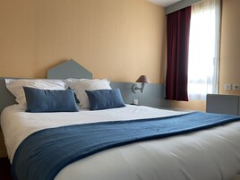 Quarto de hotel com uma cama grande, cabeceira, luminária de cabeceira e uma janela com cortinas.