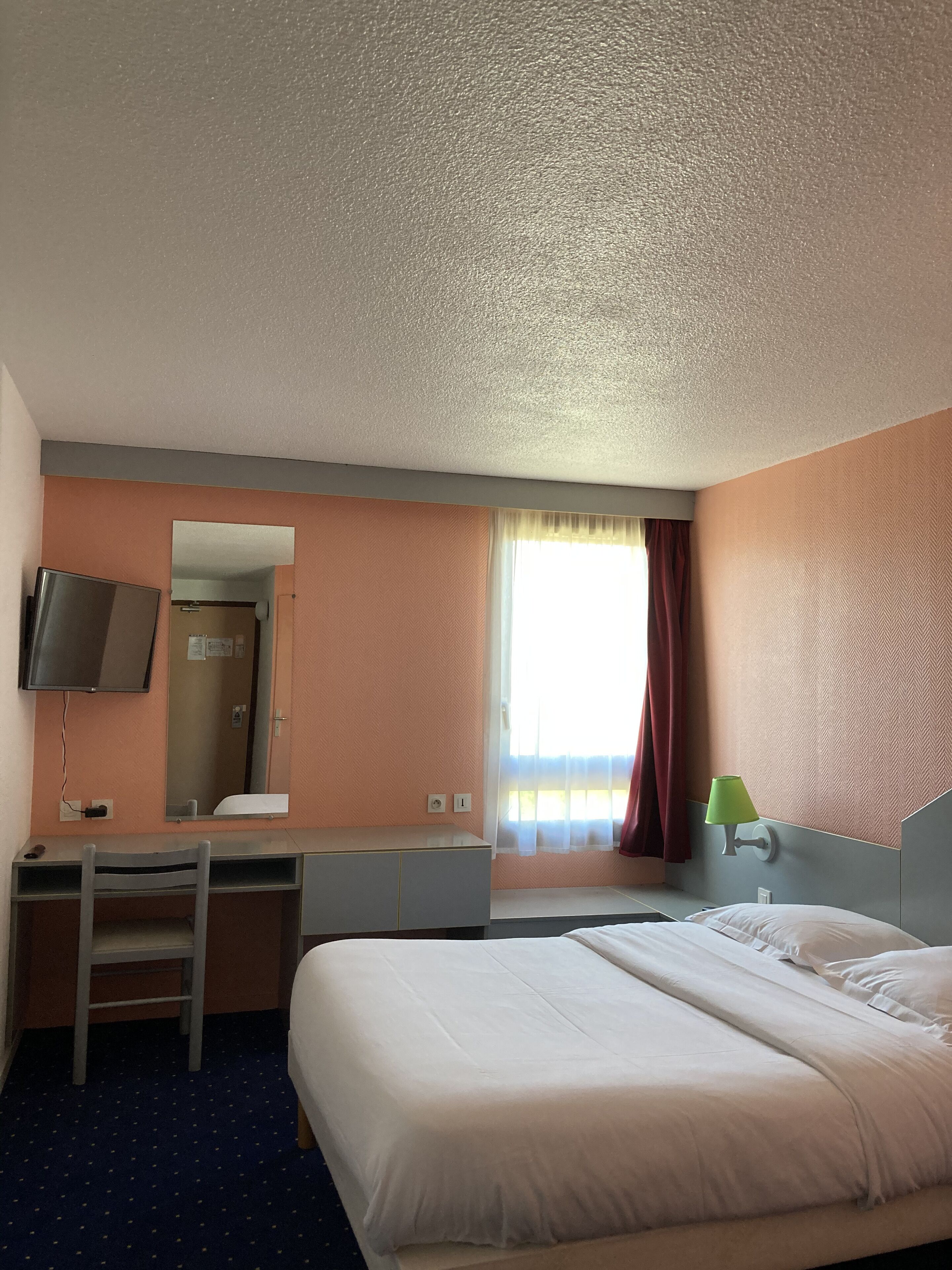 Habitación de hotel con cama, escritorio, silla, televisor y ventana con cortinas.