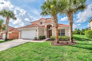 A two-story house with a red-tiled roof, a garage, and a well-maintained lawn with palm trees.