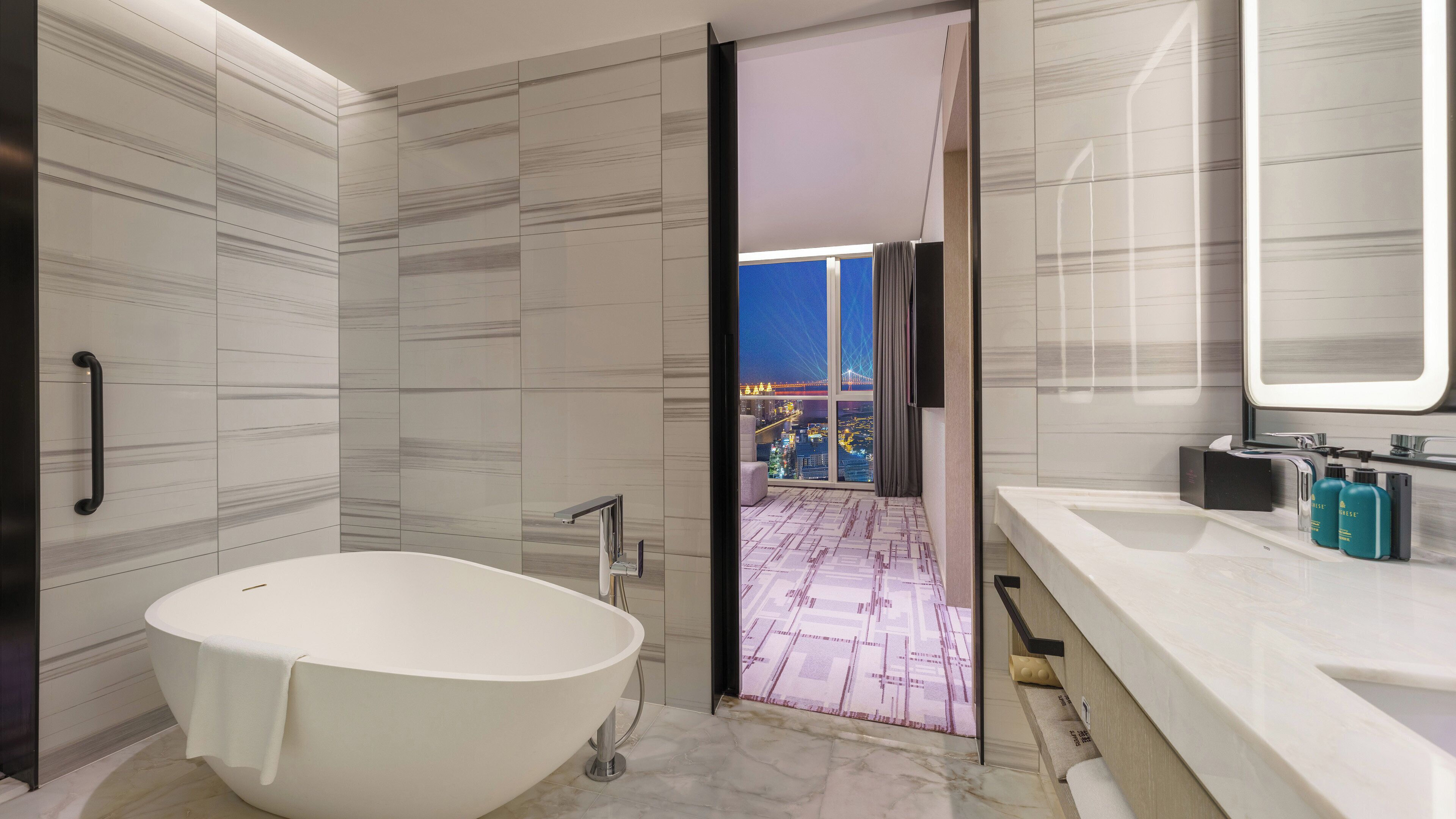 A modern bathroom with a freestanding bathtub, a large mirror, and a view of a cityscape at night.