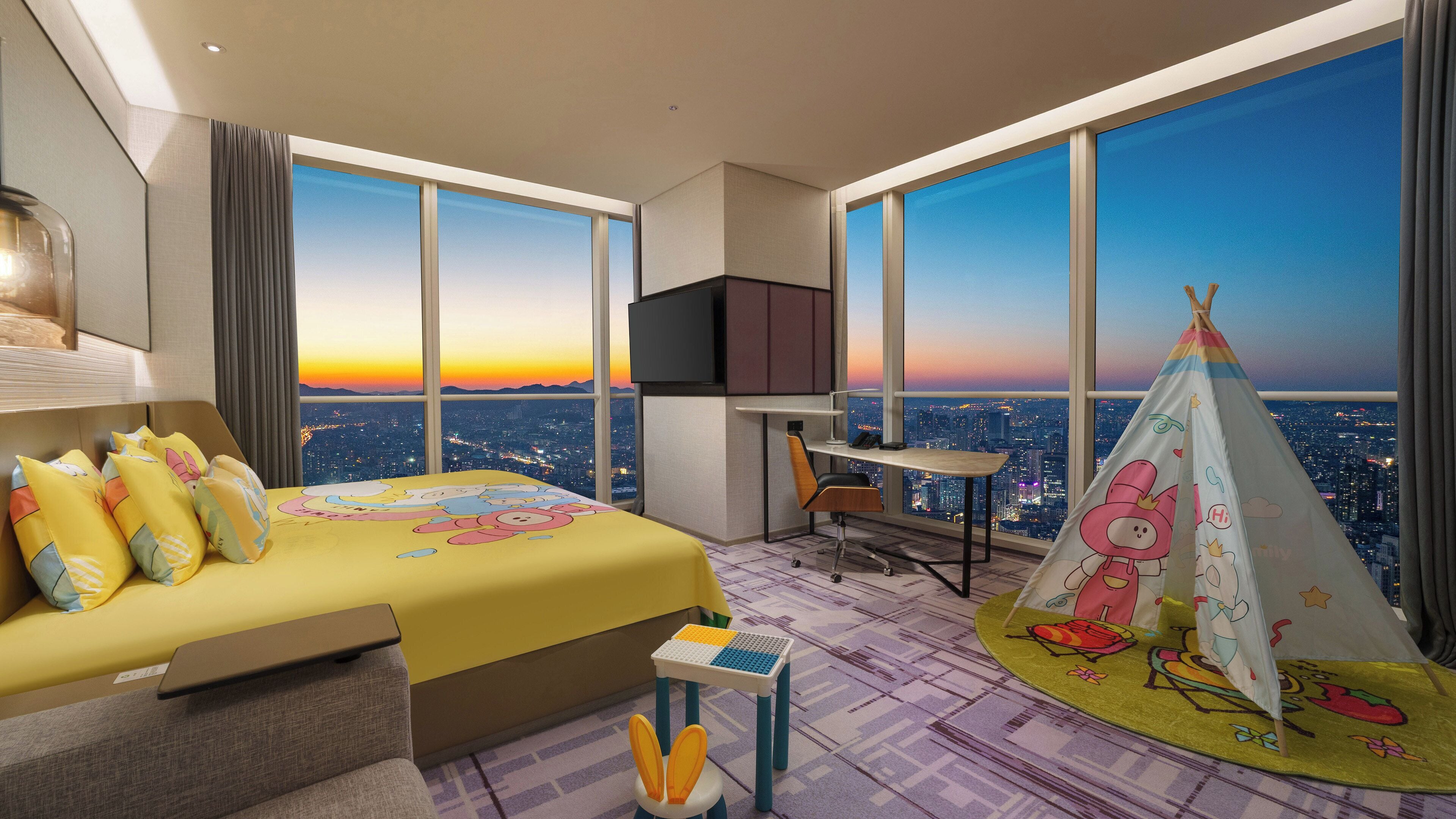 A hotel room with a large bed, a desk, a chair, a TV, and a view of the city at sunset.