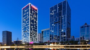 A high-rise building with a prominent sign at the top, surrounded by other modern buildings and a body of water reflecting lights. - Crowne Plaza Dalian Xinghai by IHG (Dalian)