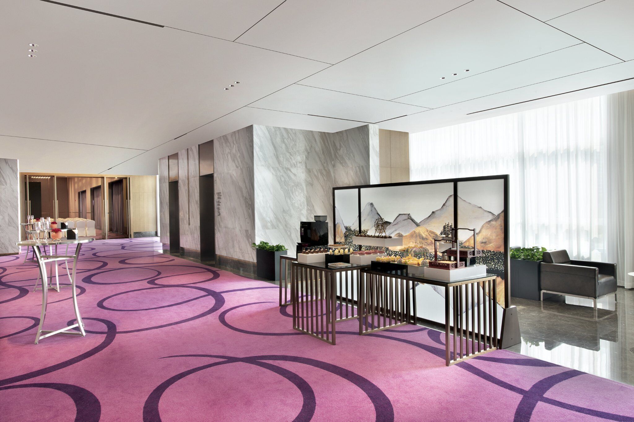 A modern hotel lobby with a purple carpet, a reception desk, and a large wall painting.