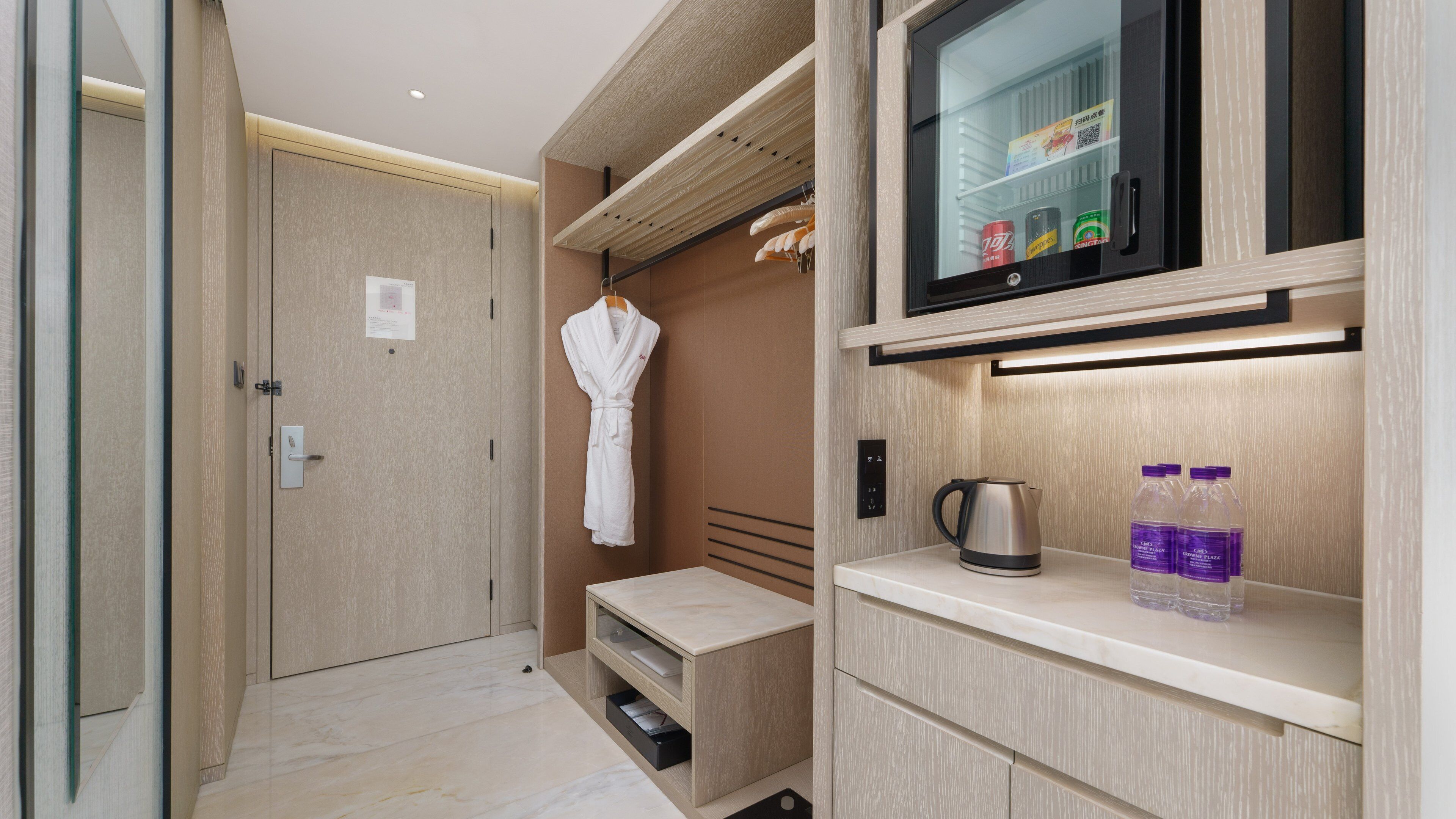 A hotel room with a minibar, a robe hanging, a kettle, and bottled water.