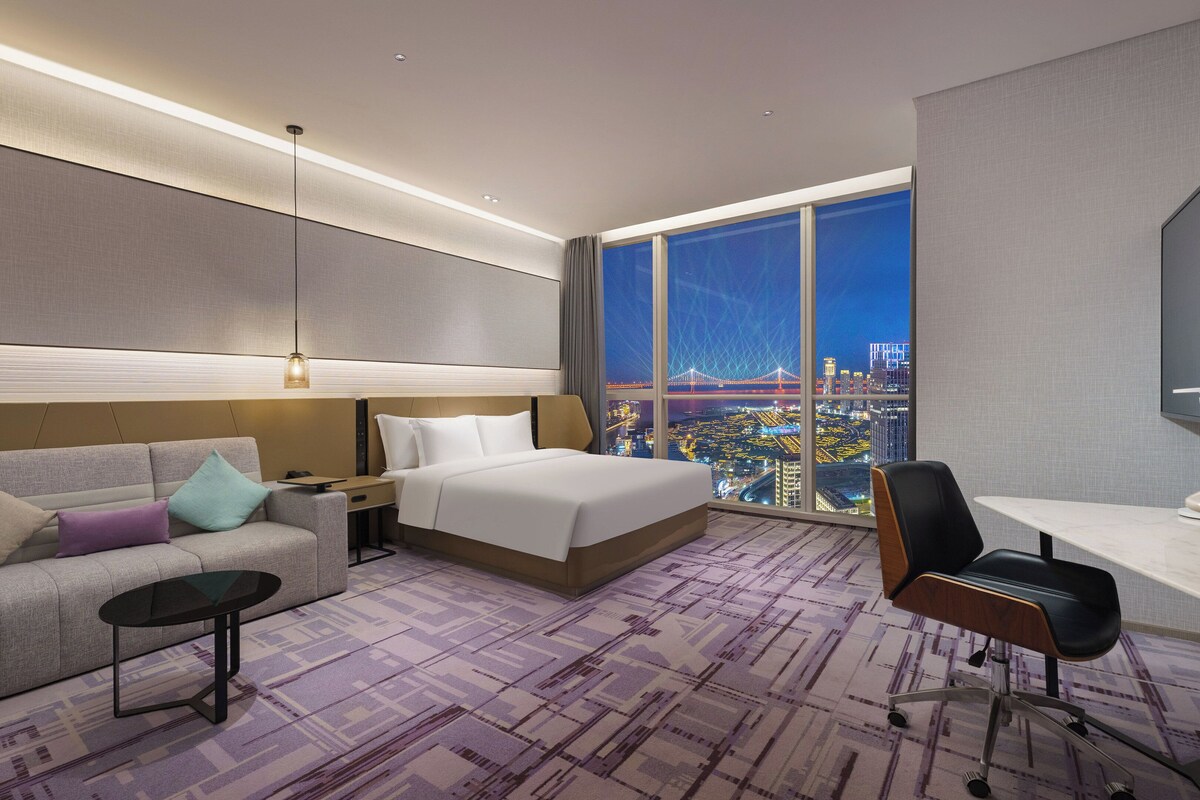 A modern hotel room with a large bed, a sofa, a desk, and a view of a cityscape.