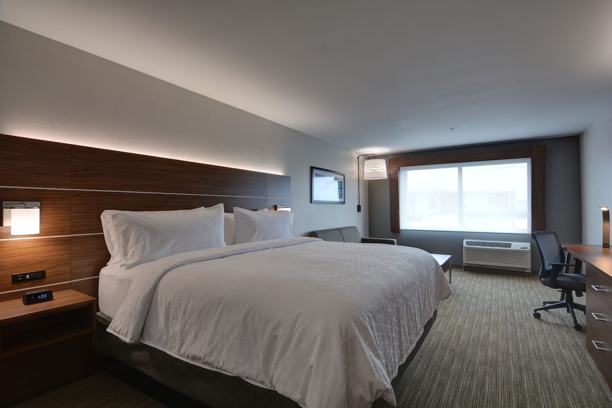 A hotel room with a large bed, a desk, a chair, a nightstand, a wall-mounted air conditioner, and a window with a view.