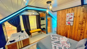 A cozy room with a wooden ceiling, a bench, a bed with towels, a bathroom with a sink, and a view of the outdoors.