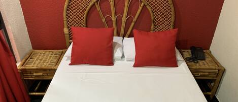 A hotel room with a bed, two red pillows, a white bedspread, a red curtain, a wicker nightstand with a phone, and a towel with the hotel name.