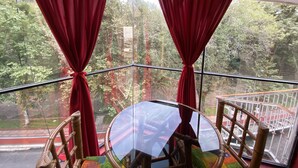 Habitación de esquina con una mesa y sillas de vidrio, cortinas rojas y vistas a árboles y un edificio rojo en el exterior.