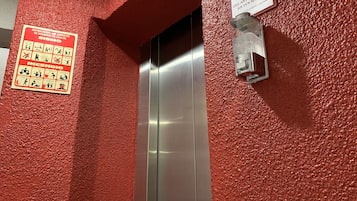 Un ascensor con interior metálico, paredes texturizadas rojas y señales de seguridad, incluyendo una de prevención de incendios y otra para la sanitización de manos.