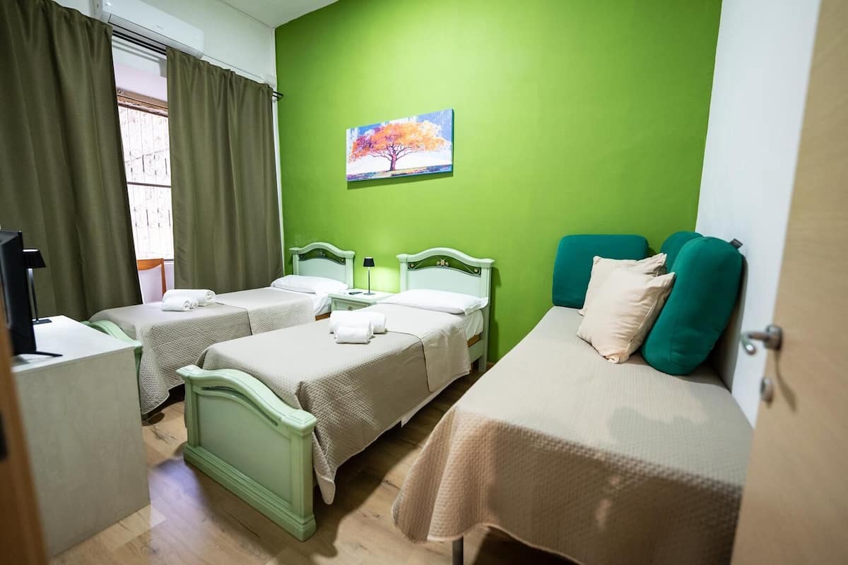 A room with two single beds, a larger bed, a painting of a tree, and a window with curtains.