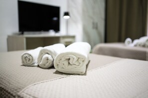 A neatly made bed with rolled white towels, a television on a stand, and a lamp in the background. - Home Josèphine (Caserta)