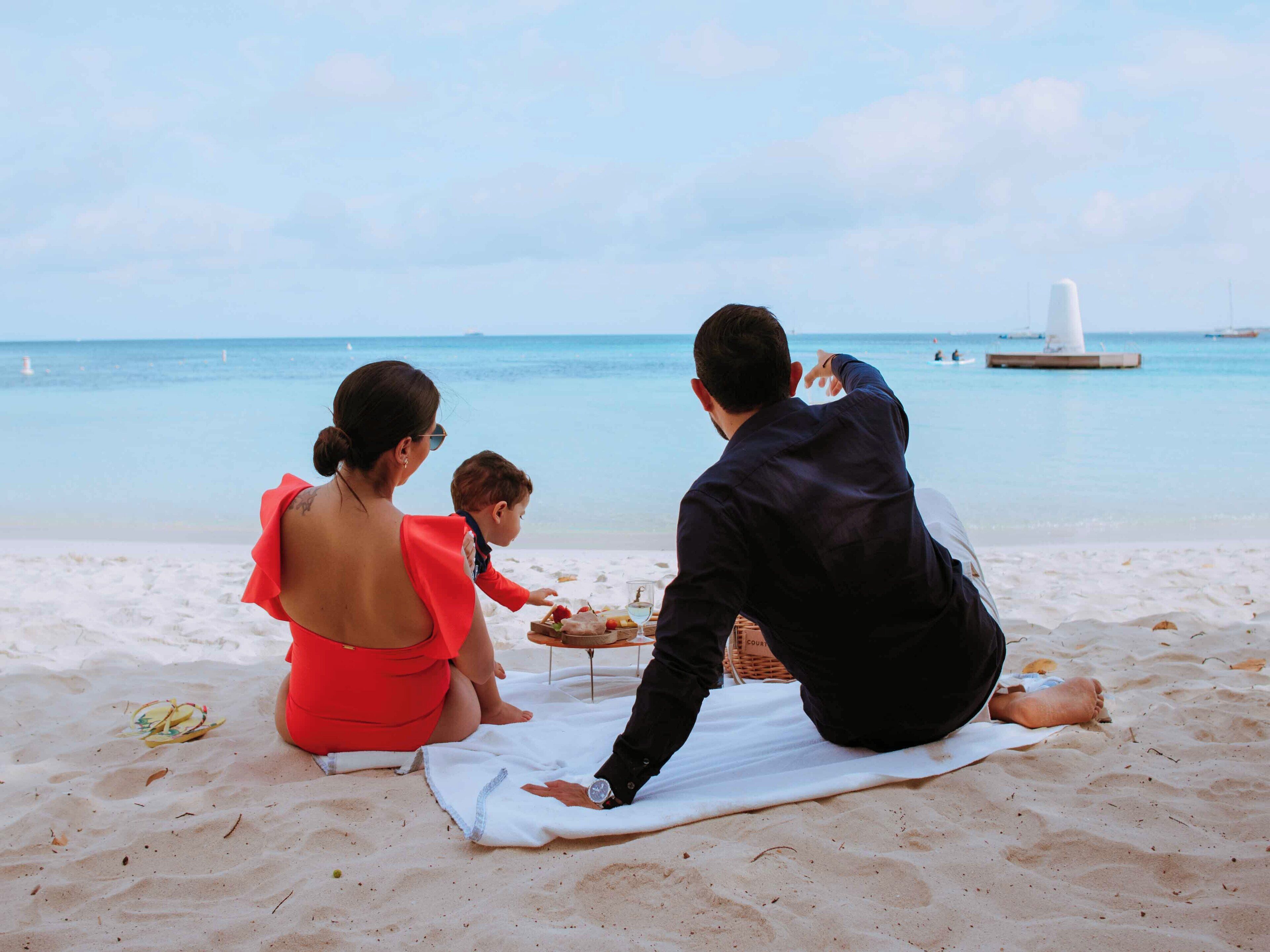 A family of three on a beach with a white umbrella, a white beach chair, and a white table with food.
