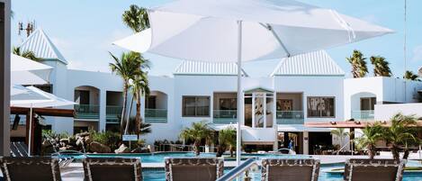 Un área de piscina con sillones de descanso y sombrillas, cielo despejado y un edificio con balcones y palmeras.