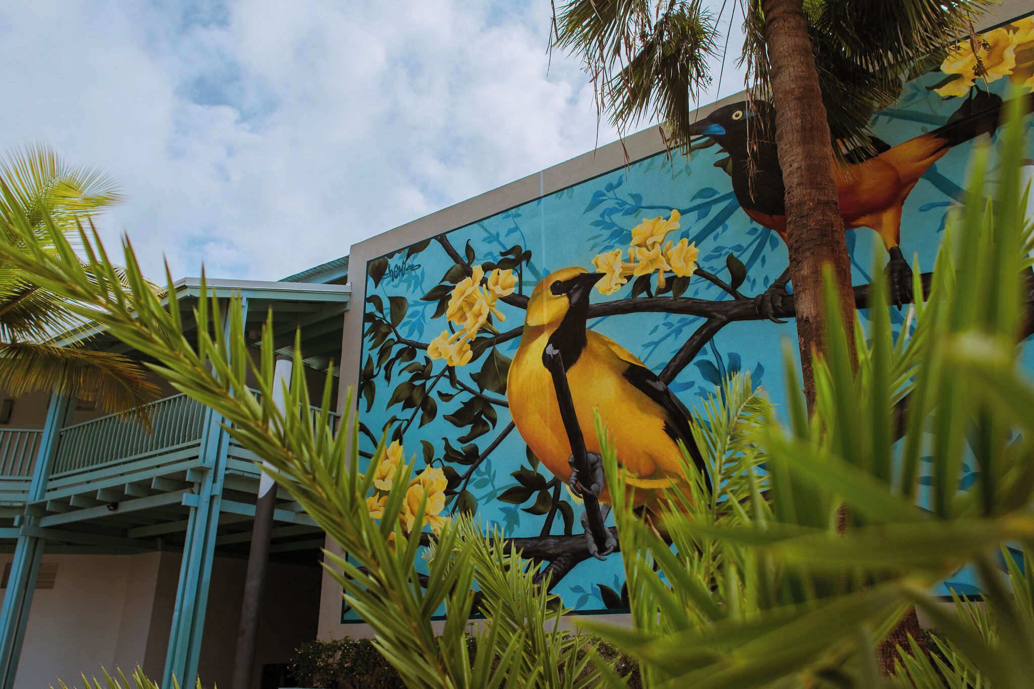 A mural of a bird on a branch with yellow flowers, a blue railing, and palm trees.