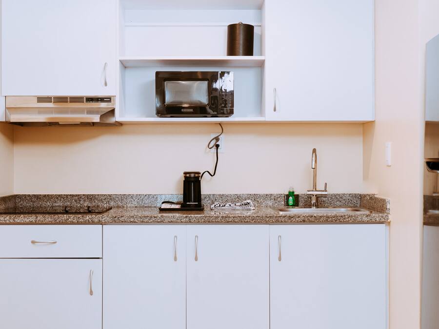 Una cocina con armarios blancos, microondas, cafetera y lavamanos.