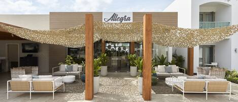A modern outdoor seating area with a canopy, a sign reading "Allegra," and a building entrance.
