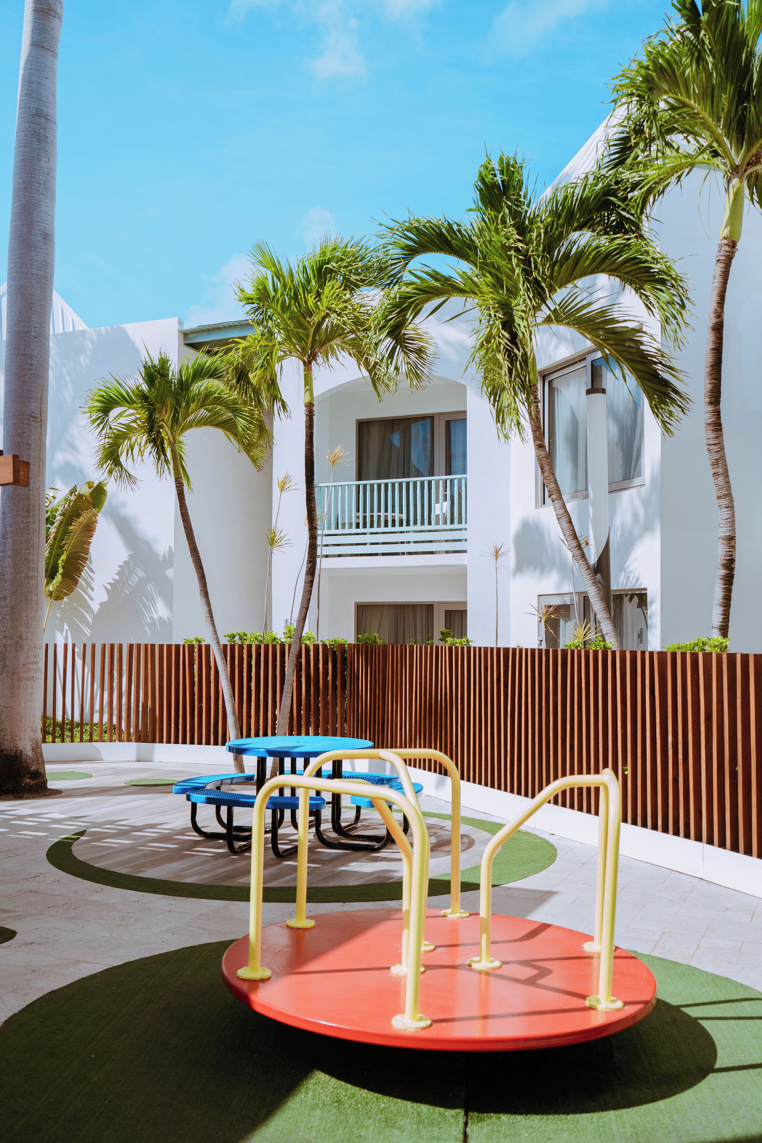 Een speeltuin met een draaiplatform, palmbomen en een gebouw met balkon op de achtergrond.