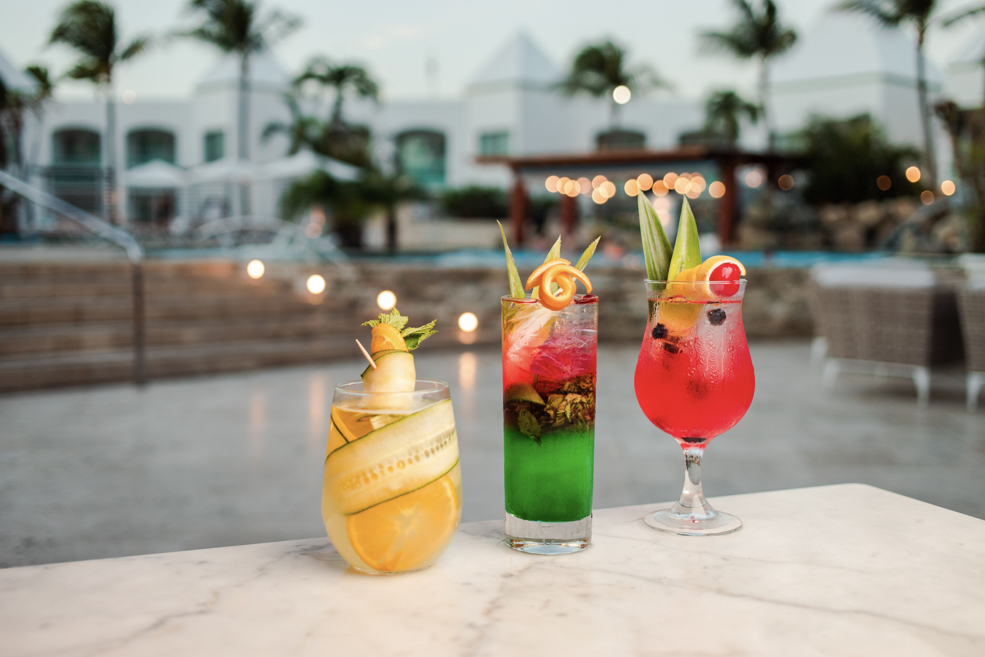 Three cocktails on a marble table with a pool and palm trees in the background.