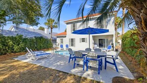 A pool area with lounge chairs and a table, surrounded by palm trees and a white building with a red roof. - Private Pool, 6 passenger Golf Cart, and 4 bikes! EV charger on site! (Destin)