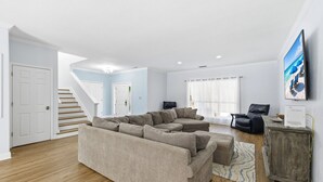 A spacious living room with a beige sectional sofa, a wooden floor, a staircase, and a flat-screen TV mounted on the wall. - Private Pool, 6 passenger Golf Cart, and 4 bikes! EV charger on site! (Destin)