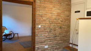 Una habitación con pared de ladrillo, una puerta de madera, una mesita con una silla y un florero con flores sobre una estantería.