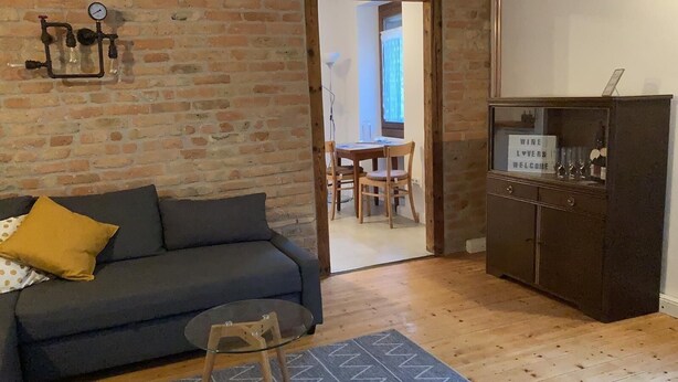 Una sala de estar acogedora con pared de ladrillo, un sofá oscuro, una mesa de centro de cristal y un mueble de madera con copas de vino.