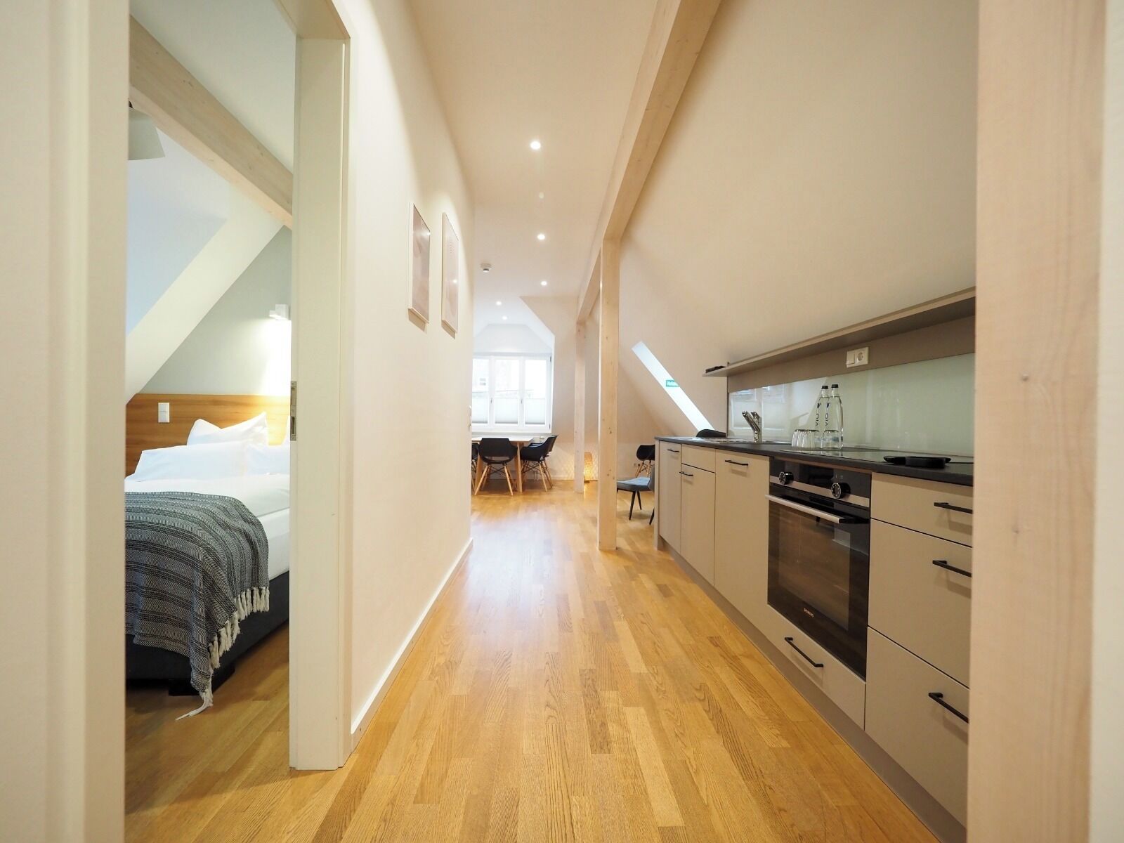 Una cocina moderna con suelo de madera, un comedor con sillas y un dormitorio con cama y ventana.