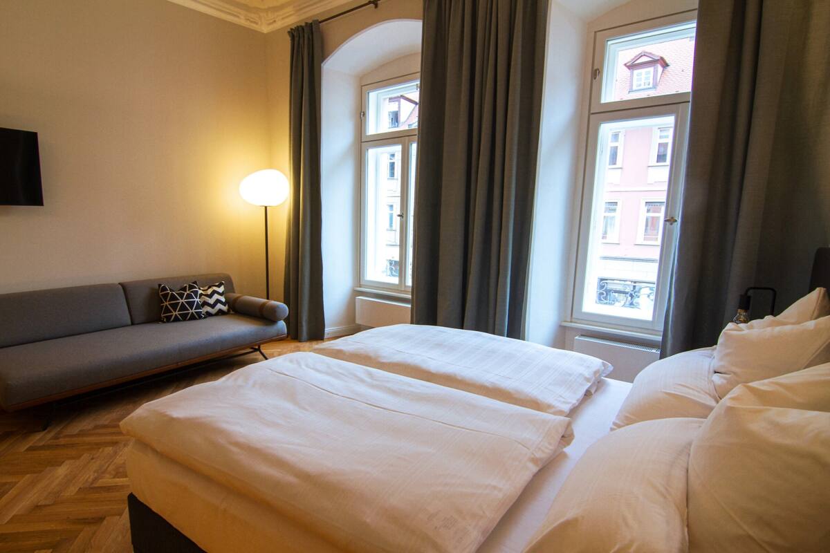 Una habitación de hotel con una cama, un sofá, un televisor y un ventanal con cortinas.