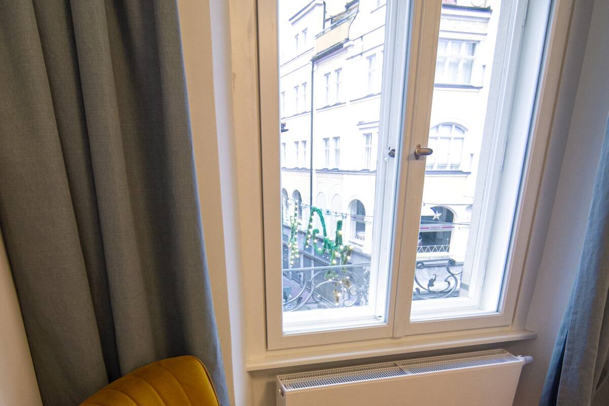 Habitación de esquina con un sillón amarillo, un radiador y una ventana que da a edificios.