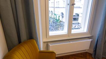 A corner room with a yellow armchair, a radiator, and a window with a view of buildings.
