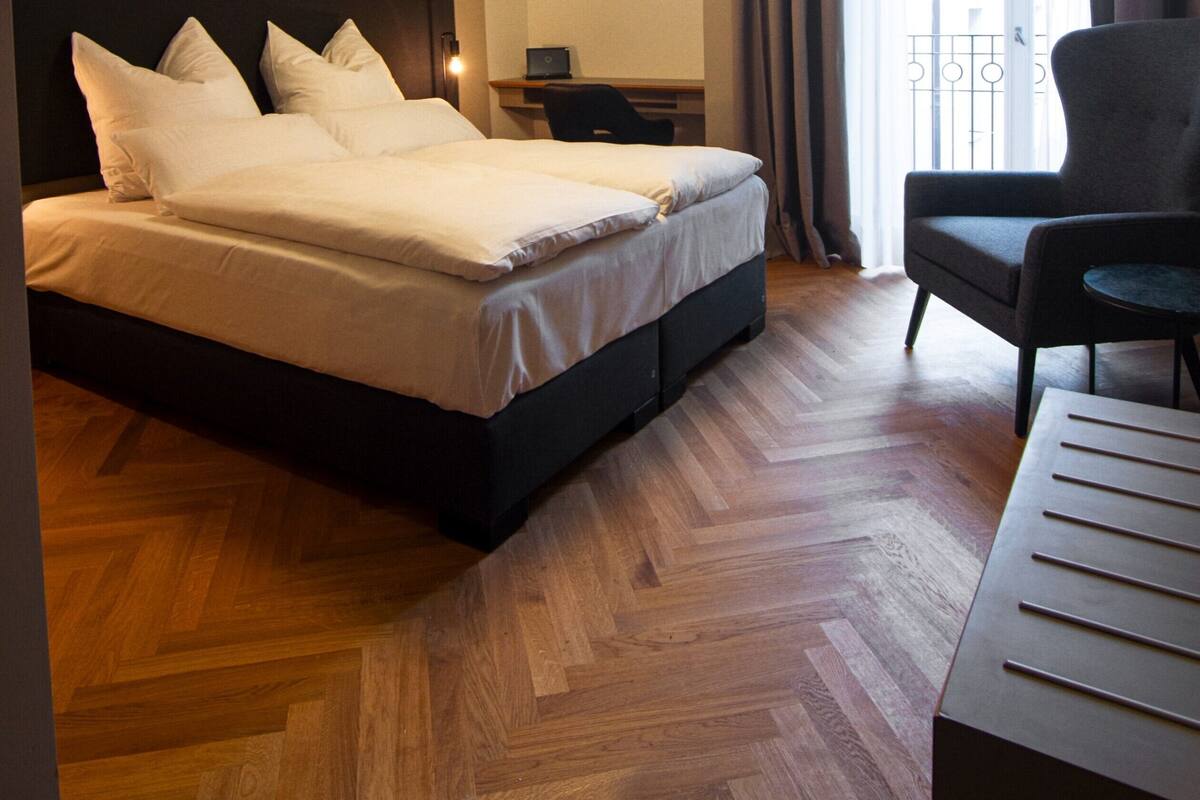 Habitación de hotel con cama, escritorio, silla, mesita de noche y un amplio ventanal con cortinas.