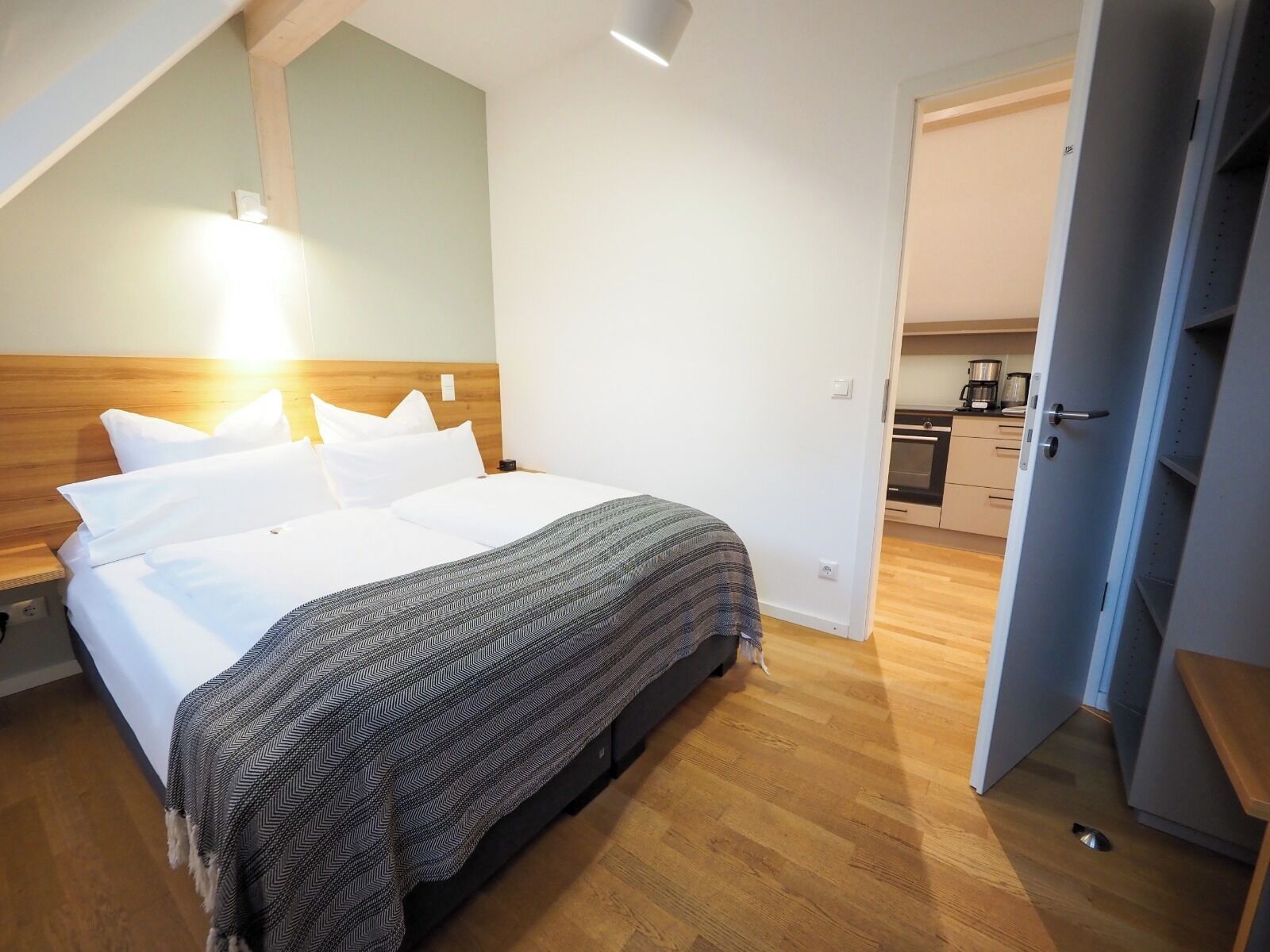 Une chambre à coucher comprenant un lit, une table de chevet, une commode, une couverture à motifs, une tête de lit en bois et une vue sur un coin cuisine.