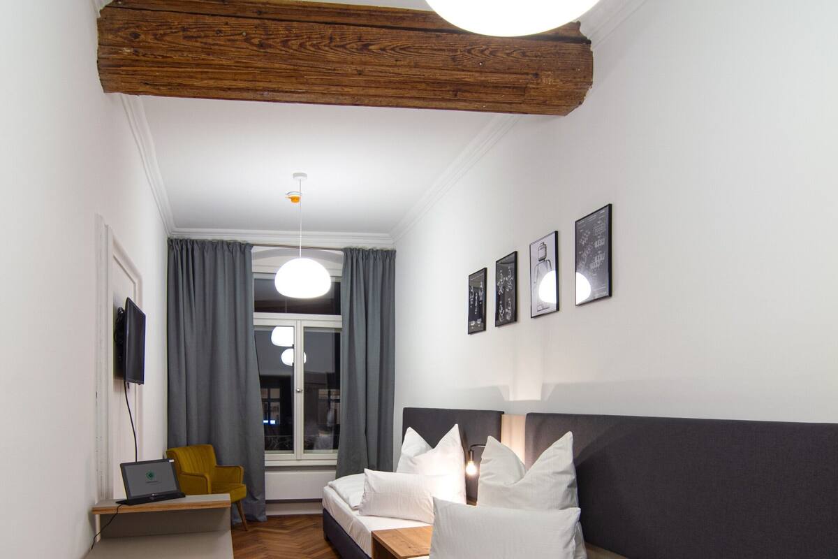 Una habitación de hotel moderna con suelo de madera, sofá, cama, escritorio con una laptop y televisor en la pared.
