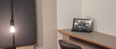 Una habitación pequeña y moderna con un escritorio de madera, una silla negra, una lámpara de escritorio y un marco digital con fotografías en la pared.