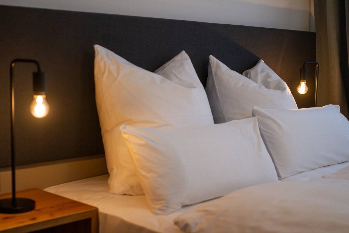 Una cama bien hecha con sábanas y almohadas blancas, una mesita de noche de madera con una lámpara, y un cabecero oscuro.