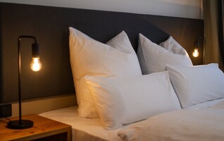 Una cama bien hecha con sábanas y almohadas blancas, una mesita de noche de madera con una lámpara, y un cabecero oscuro.