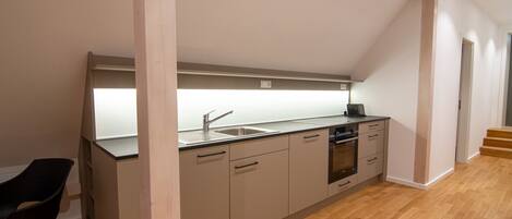 A modern kitchen with a dishwasher, oven, and sink. There is a wooden floor and a black chair.