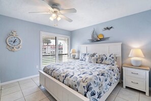 A bedroom with a bed, a ceiling fan, a clock, a window with blinds, and a nightstand with a lamp.