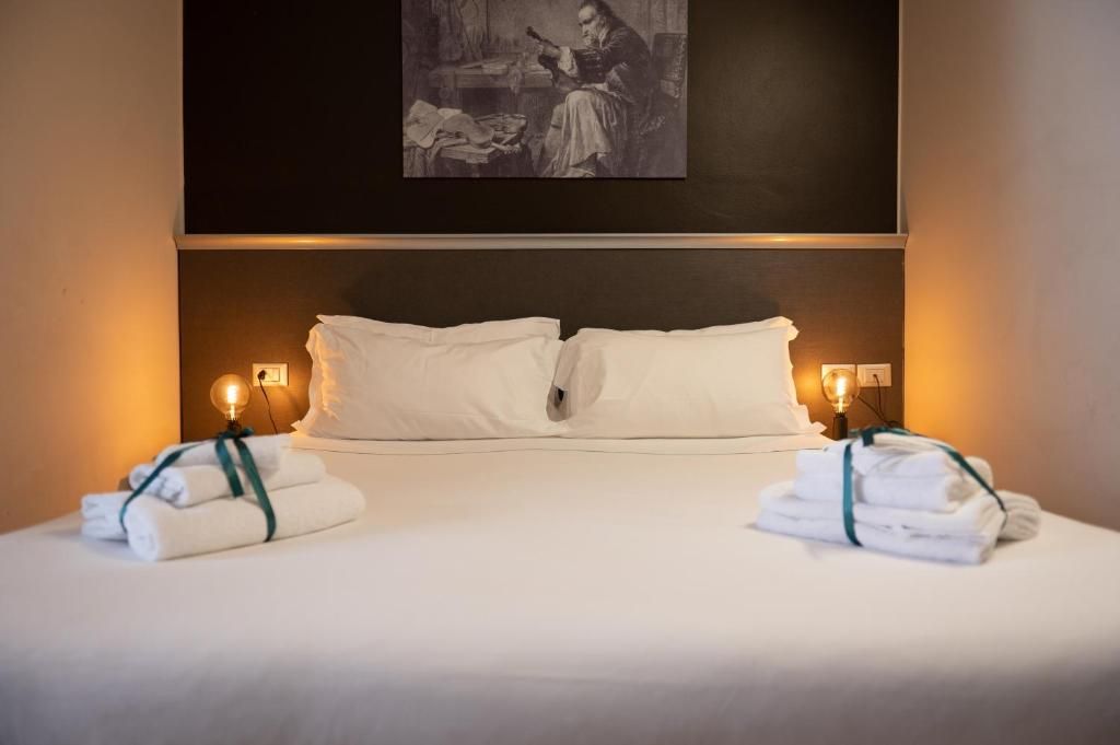 Een netjes opgemaakt bed met witte lakens en twee handdoeken aan groene linten, een schilderij aan de muur boven het bed, en bedlampen.