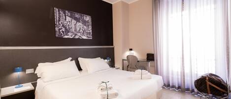 A hotel room with a large bed, a desk with a chair, a lamp, and a window with sheer curtains.