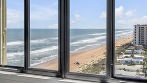 A view from a window overlooking a beach with ocean waves, a sandy shore, and buildings in the distance. - Luxury direct oceanfront 3/3 corner penthouse with heated pool and garage parking (Ormond Beach)