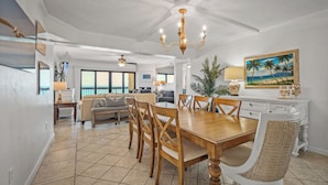 A spacious living area with a dining table, chairs, a sofa, and a large window. - Luxury direct oceanfront 3/3 corner penthouse with heated pool and garage parking (Ormond Beach)