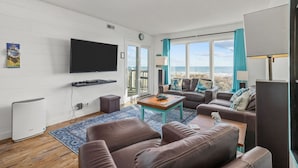 A modern living room with a flat-screen TV, leather sofas, a coffee table, and a large window with a view of the beach. - Immaculate 3/2 Oceanfront Condo near Main St, Pool (North Myrtle Beach)