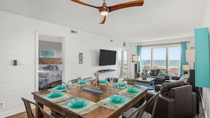 A dining area with a wooden table set for four, a ceiling fan, and a view of the ocean through large windows. - Immaculate 3/2 Oceanfront Condo near Main St, Pool (North Myrtle Beach)