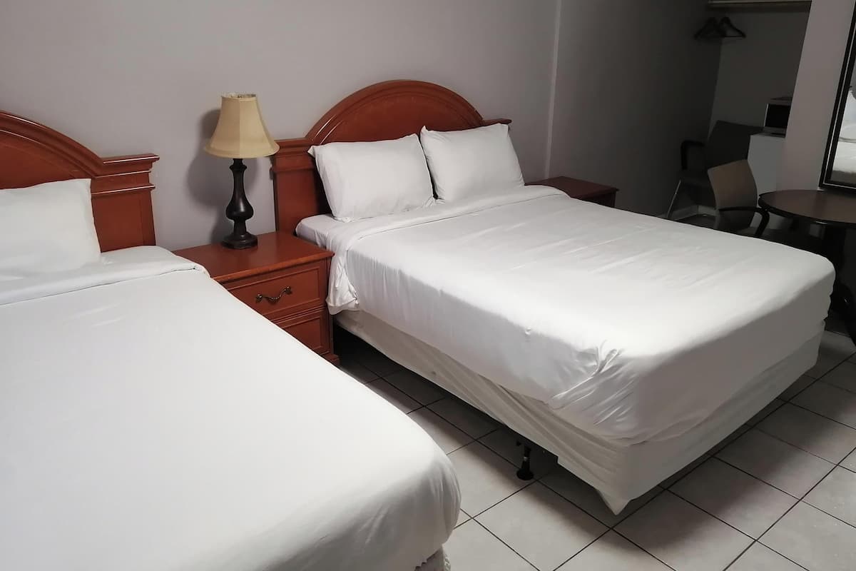 Quarto de hotel com duas camas, uma mesa de cabeceira com abajur, uma pequena escrivaninha com cadeira e um espelho na parede.
