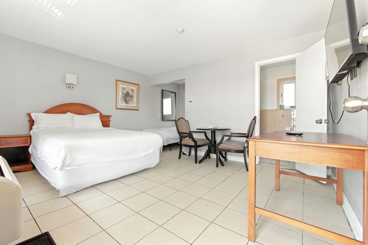Quarto de hotel com cama, escrivaninha, duas cadeiras, mesinha, espelho e televisão presa na parede.