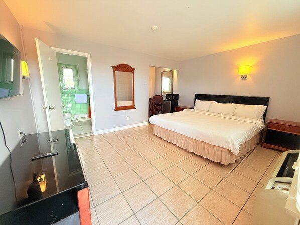 Une chambre d’hôtel avec un grand lit, un bureau avec un ordinateur, un miroir et une salle de bain visible à travers une porte ouverte.
