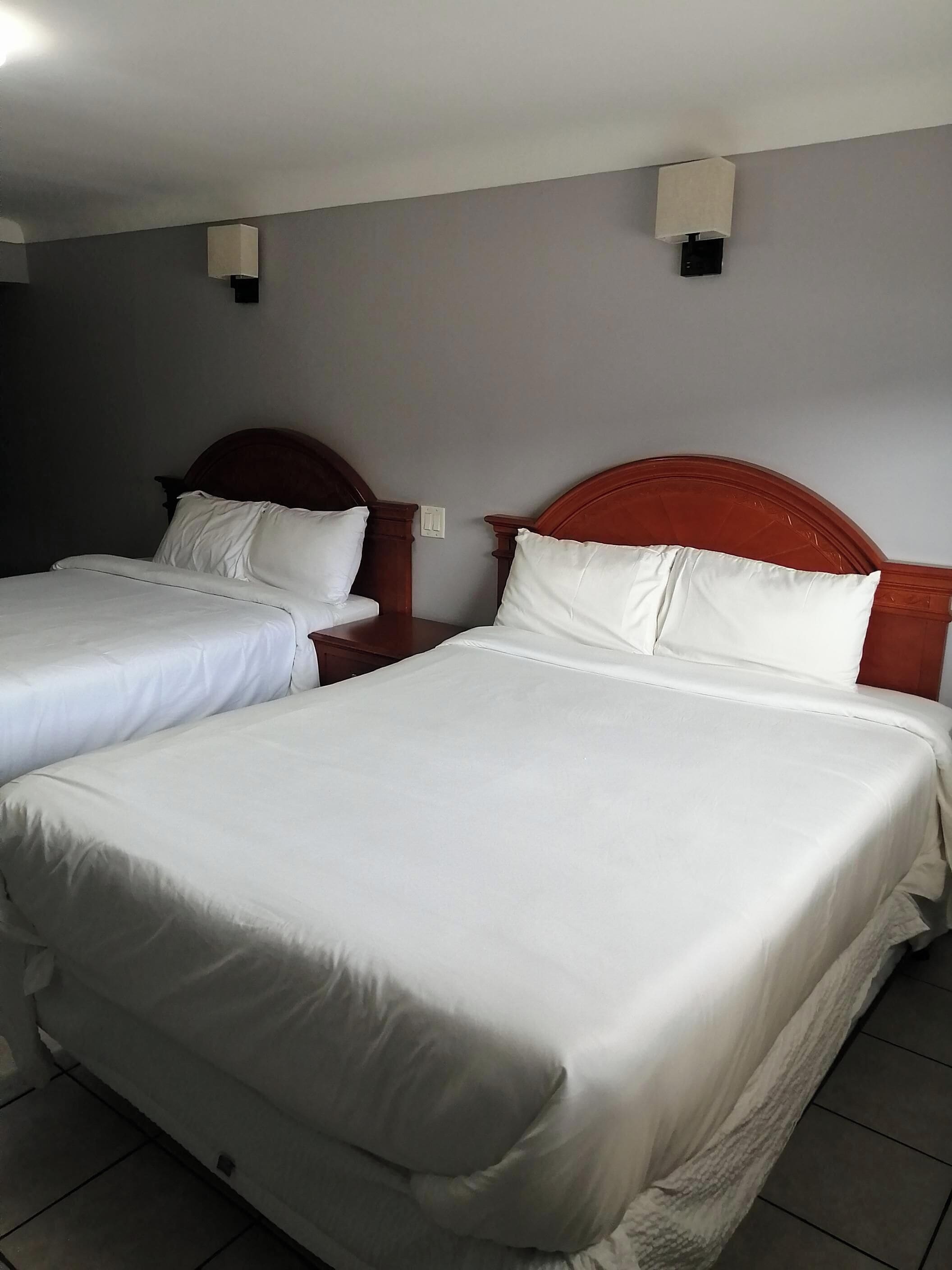 Ett hotellrum med två separata sängar, båda med vita sängkläder och sänggavlar i trä. Det finns två sängbord, det ena med en lampa och det andra med ett eluttag. Väggarna är målade i en neutral färg, och det finns en taklampa.